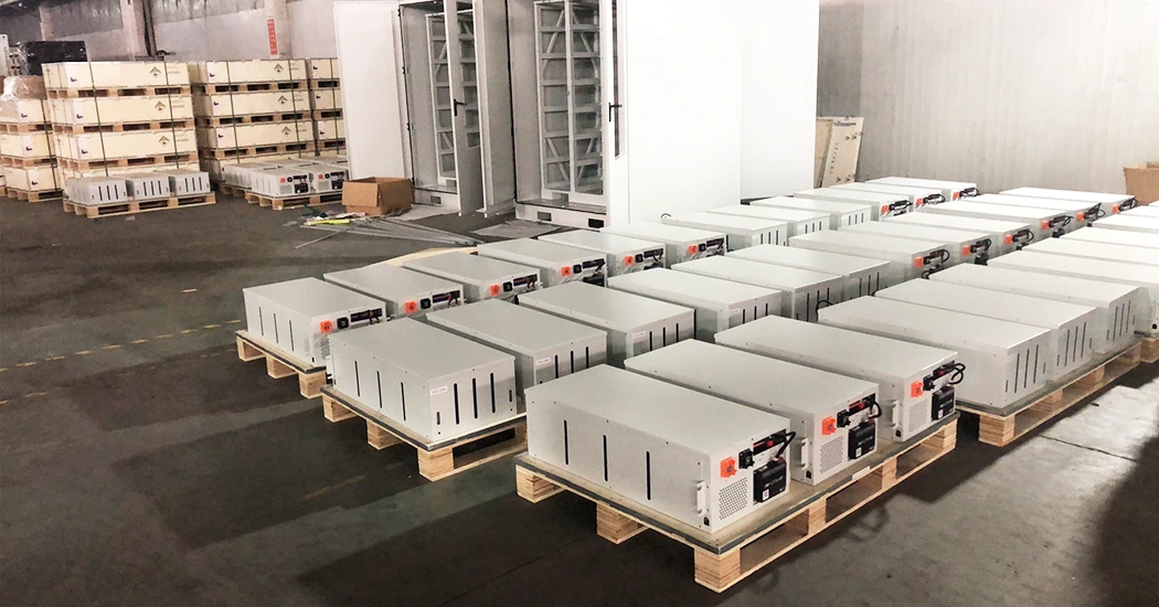 Rows of modular lithium battery units stacked on wooden pallets inside a warehouse, with battery racks and crates in the background.