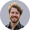A headshot of a smiling man with brown hair and a beard, wearing a plaid shirt, within a circular frame against a grey background.