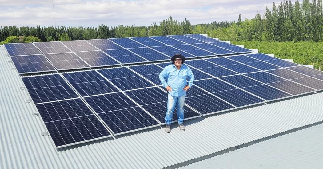 A smiling man in a blue shirt and hat standing proudly on a corrugated metal roof next to a newly installed large array of solar panels.