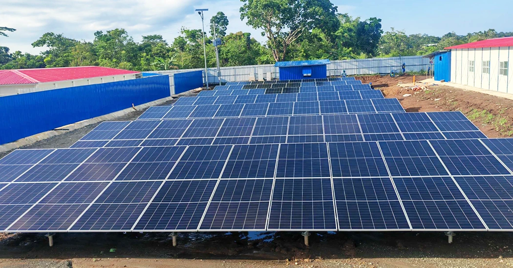 Wide front view of neatly arranged ground-mounted solar panel arrays surrounded by site enclosures and greenery.