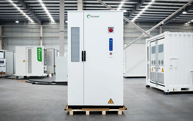 Front view of a white Sunpal commercial battery storage cabinet resting on a wooden pallet inside a manufacturing facility, featuring ventilation grilles, status lights, and an emergency stop button.