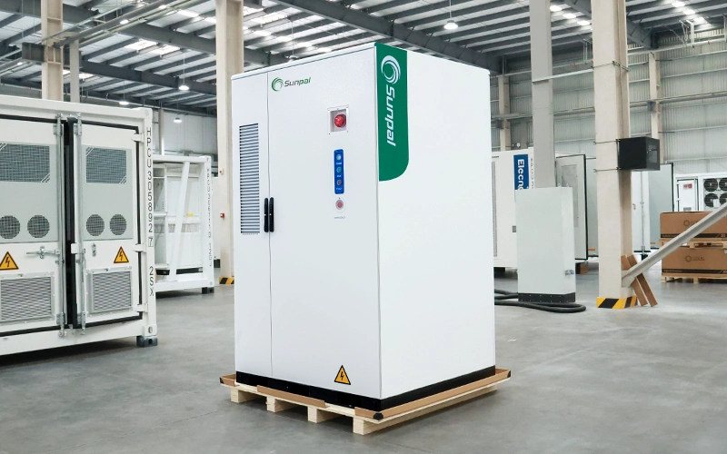 Side profile of a white Sunpal commercial energy storage cabinet sitting on a wooden pallet inside a manufacturing facility, showcasing the side ventilation, branding, and control panel.