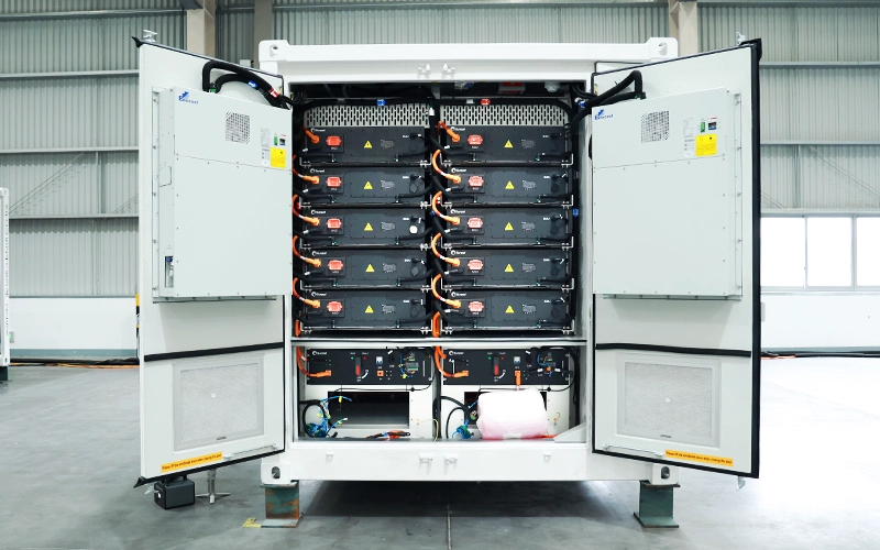 An open white energy storage container revealing two stacks of black battery modules with orange and blue cabling. It shows the main connection panels and the overall wiring layout inside a factory setting.