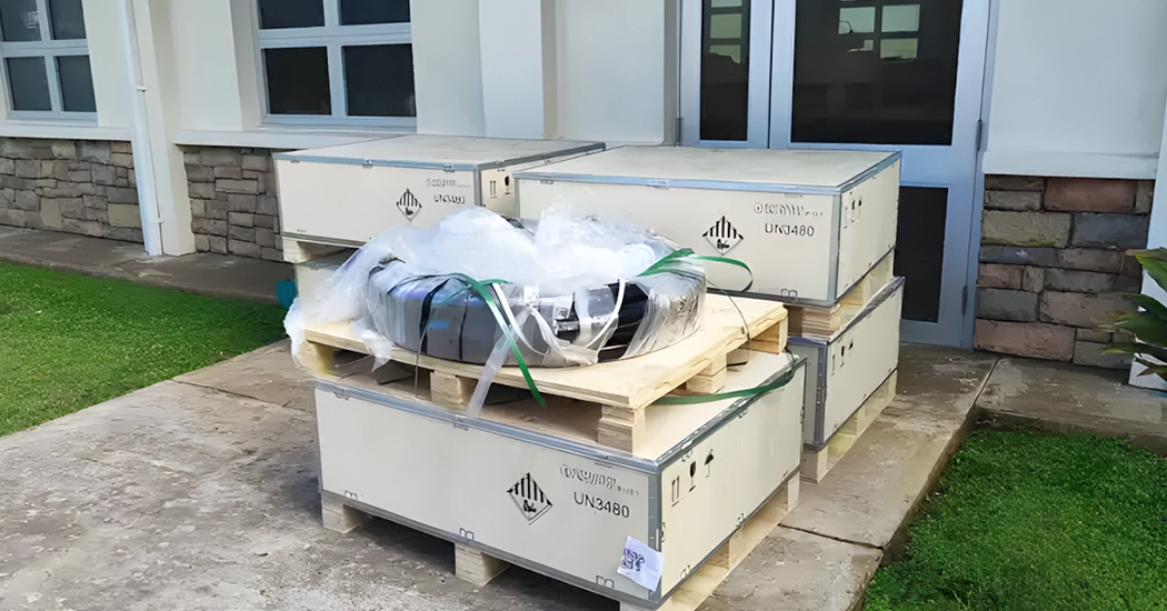 Packaged wooden crates with UN3480 lithium battery hazard labels and cabling delivered outside a facility building.