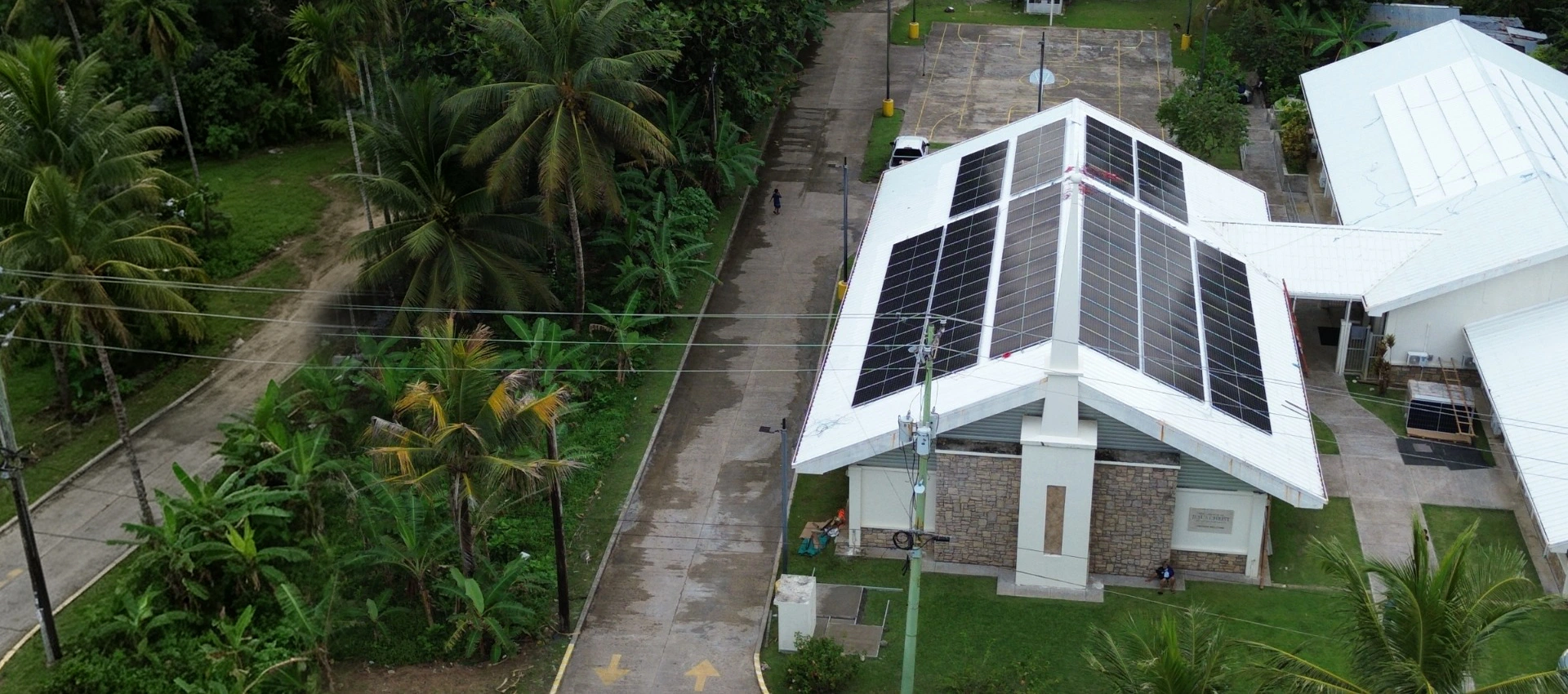 Aerial view of solar panels installed on a white rooftop in Micronesia surrounded by tropical trees.