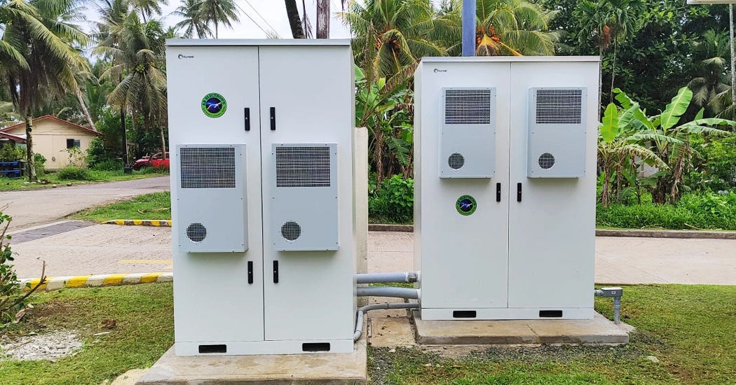 Two outdoor energy storage cabinets installed on concrete pads surrounded by tropical greenery in Micronesia.