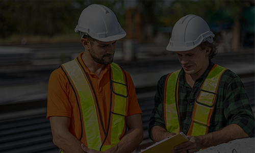 Two construction engineers in hard hats and safety vests review plans on a clipboard at an outdoor construction site.