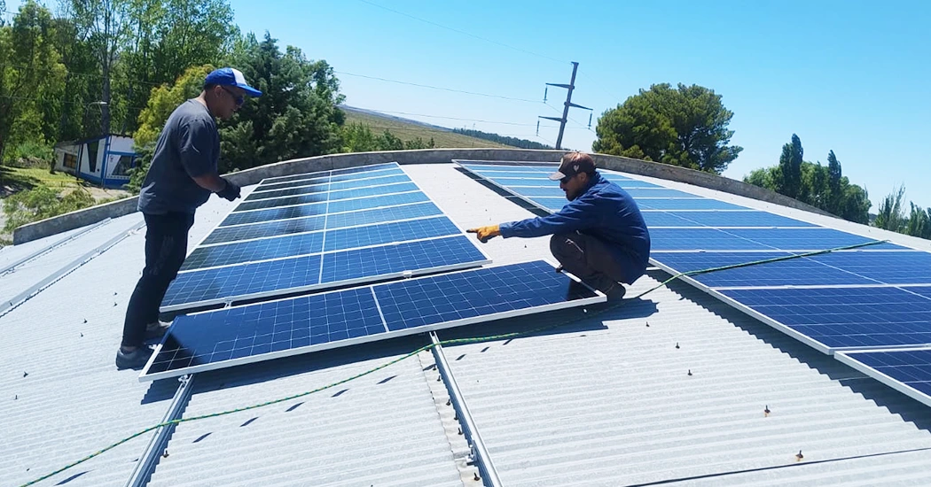Two technicians in caps inspect and install solar panel arrays on a curved corrugated metal roof under a clear blue sky.