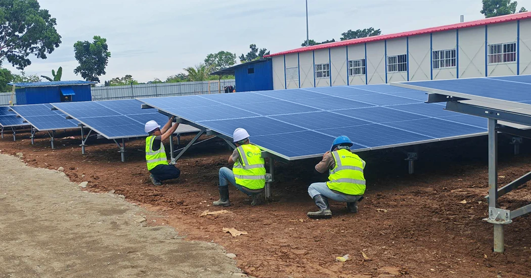 Three workers in safety gear and helmets carefully install ground-mounted solar panels at a site, crouched in a dirt area.