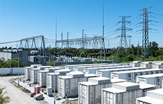Utility-scale battery energy storage power plant featuring rows of white containerized storage units deployed near high-voltage electrical transmission towers.
