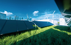 Solar panel installation on a green lawn with background wind turbines, energy storage enclosures, and a modern city skyline under a clear blue sky.